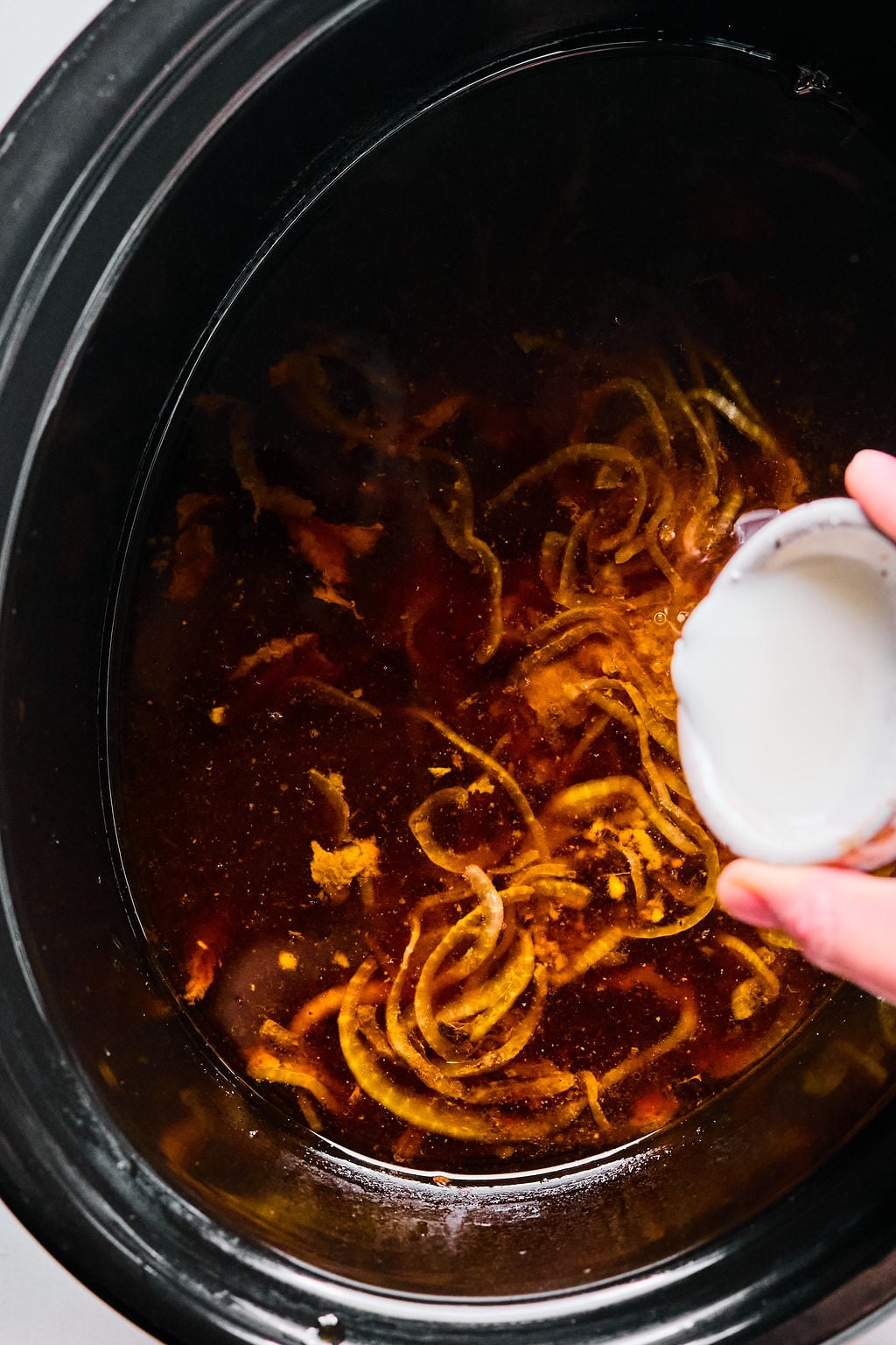 A hand pours a small bowl of cornstarch and water into a slow cooker filled with a dark broth and sliced onions.