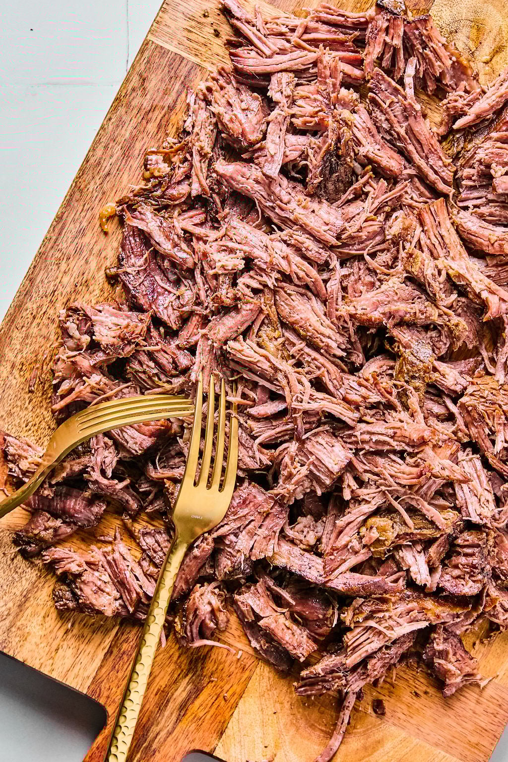 Shredded cooked beef spread on a wooden cutting board with two metal forks placed among the meat.