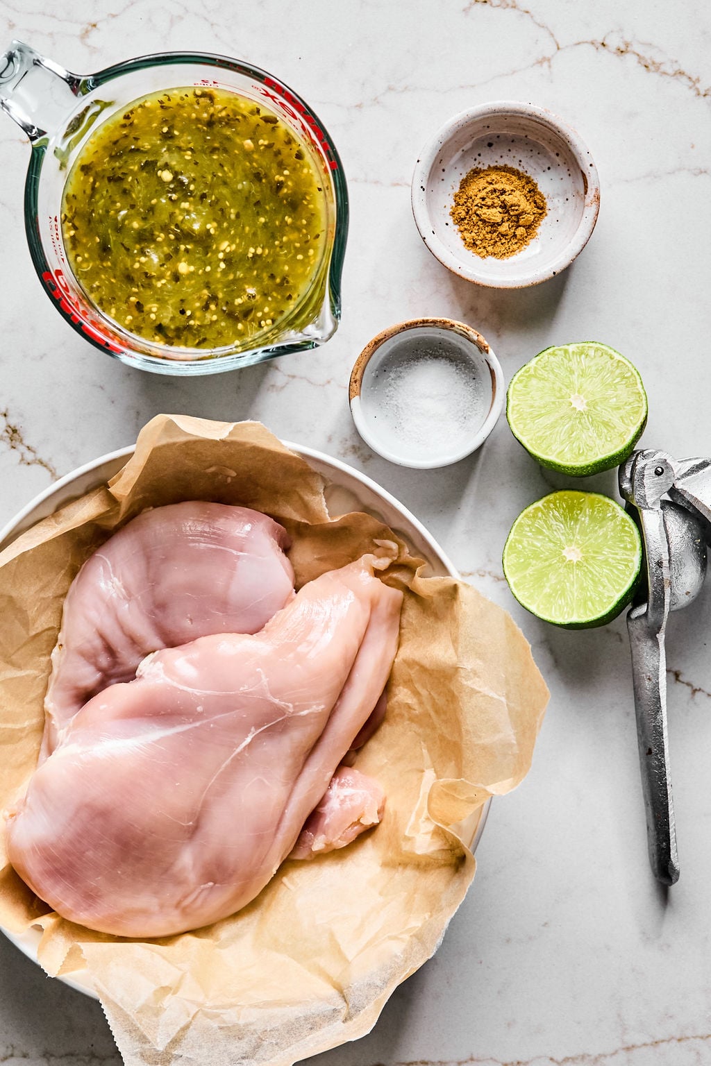 Raw chicken breasts in a bowl with parchment paper, a measuring cup of green sauce, a halved lime, a citrus squeezer, and bowls of spices and salt on a marble surface.