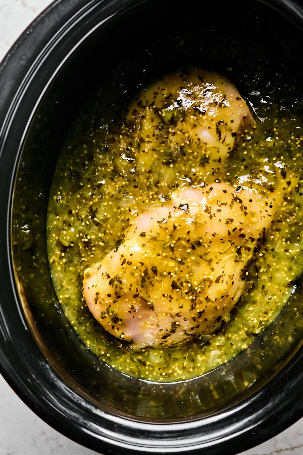 Two raw chicken breasts covered in green salsa and herbs inside a black slow cooker.