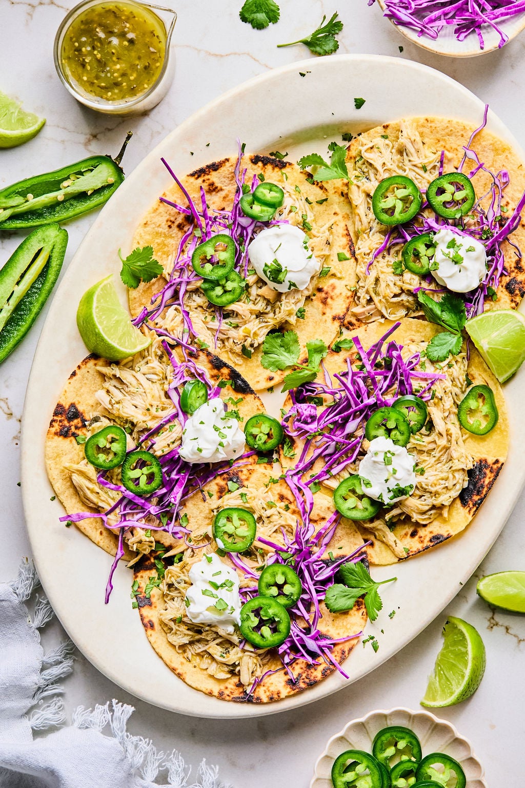 A platter of four shredded chicken tacos topped with purple cabbage, sliced jalape&ntilde;os, sour cream, cilantro, and lime wedges, with extra sauce and sliced jalape&ntilde;os on the side.