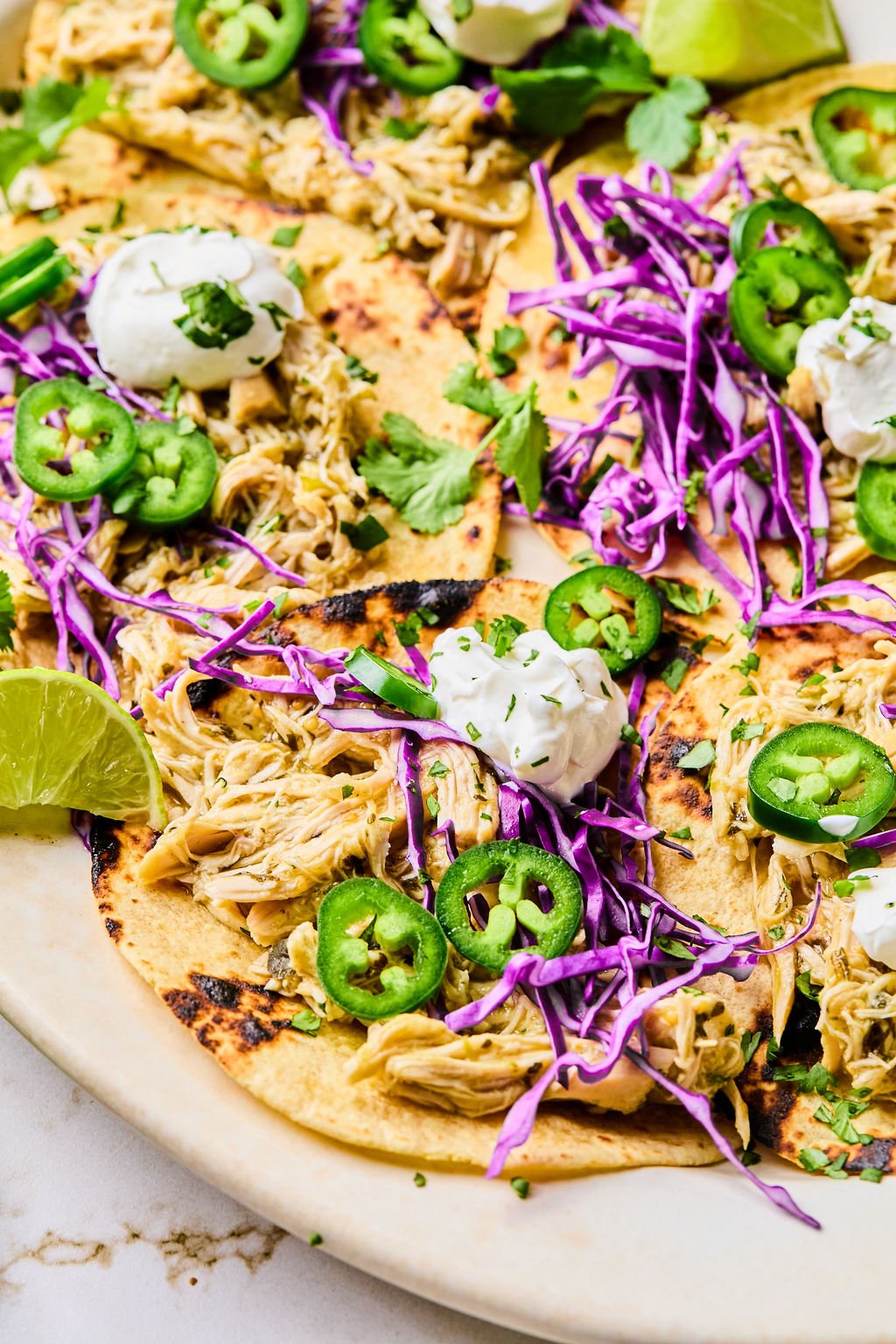 Three shredded chicken tacos on corn tortillas topped with sliced jalape&ntilde;os, shredded purple cabbage, cilantro, and sour cream.