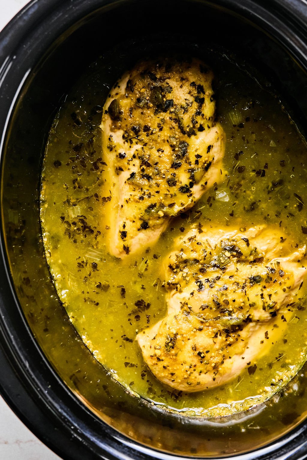 Two seasoned chicken breasts cooking in a slow cooker with a yellow, herb-infused sauce.