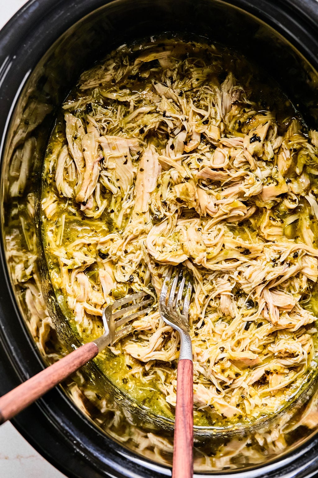 Shredded chicken in sauce inside a slow cooker, with two forks used for pulling the meat apart.