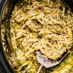 Shredded chicken mixed with green sauce in a slow cooker, with a metal serving spatula resting inside.