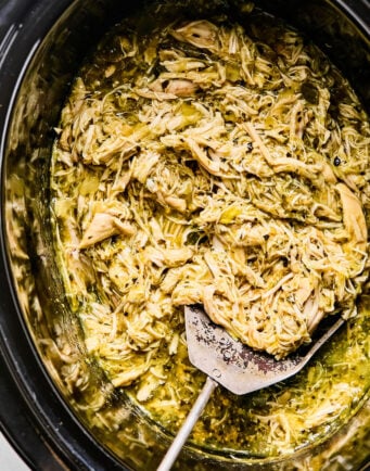 Shredded chicken mixed with green sauce in a slow cooker, with a metal serving spatula resting inside.