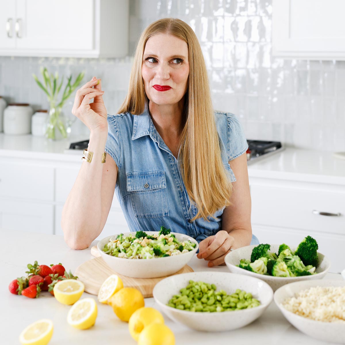 A woman with long blonde hair sits at a kitchen counter with bowls of salad, broccoli, edamame, strawberries, and lemons in front of her.