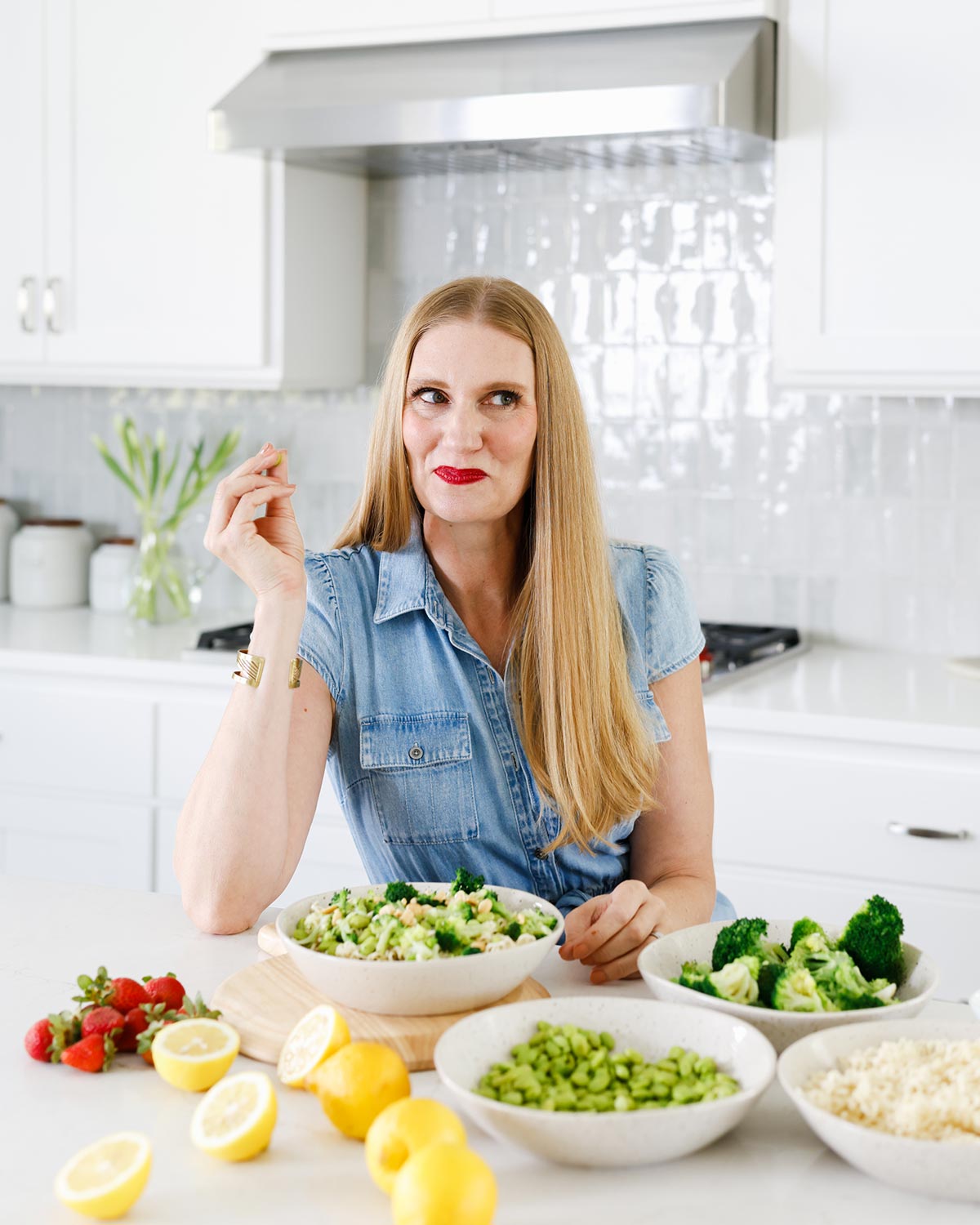 Woman in a denim shirt sits at a kitchen counter with bowls of fresh vegetables, salad, strawberries, and lemons in front of her.