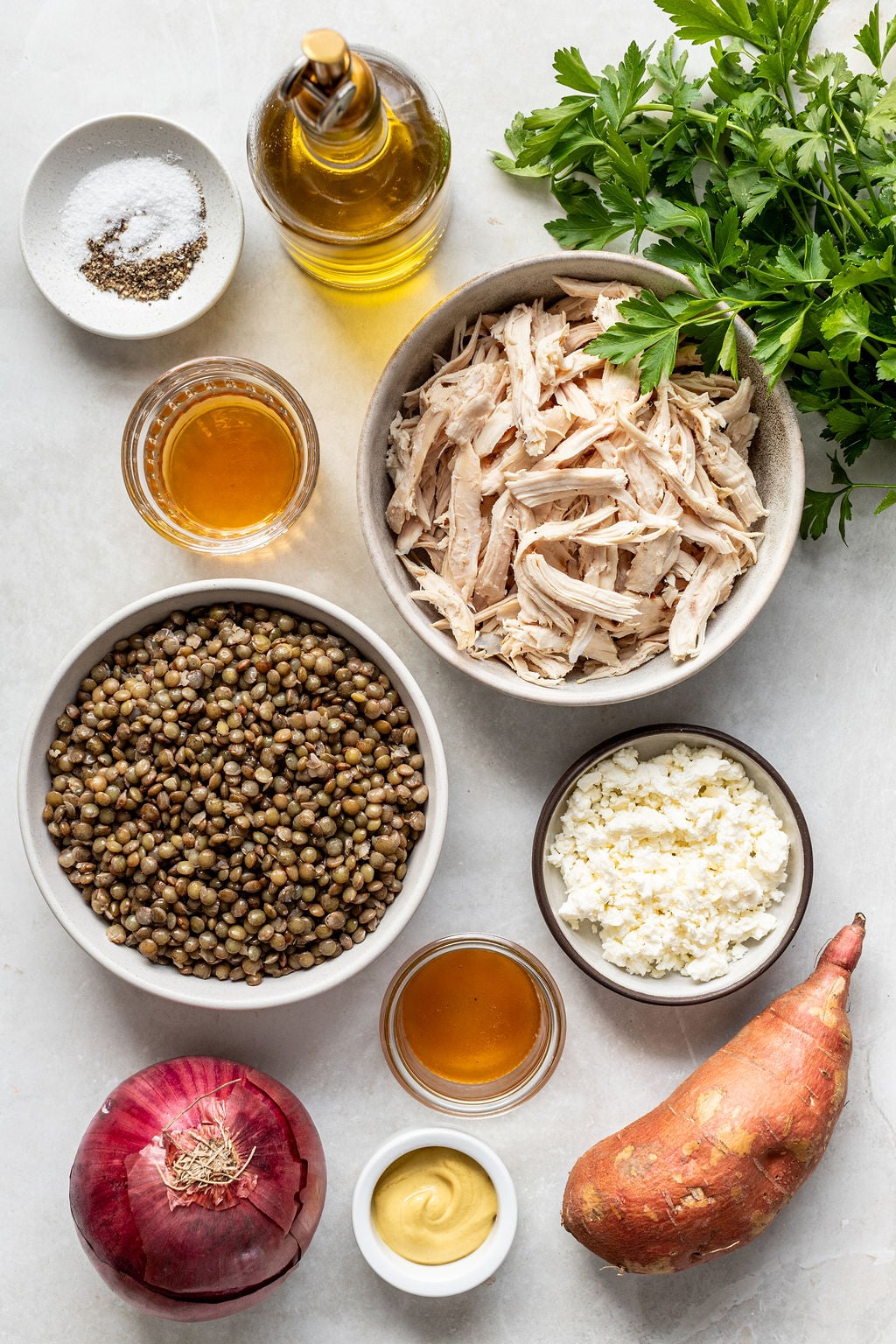 Various ingredients including shredded chicken, cooked lentils, cheese, red onion, sweet potato, olive oil, salt, pepper, and fresh parsley arranged on a countertop.
