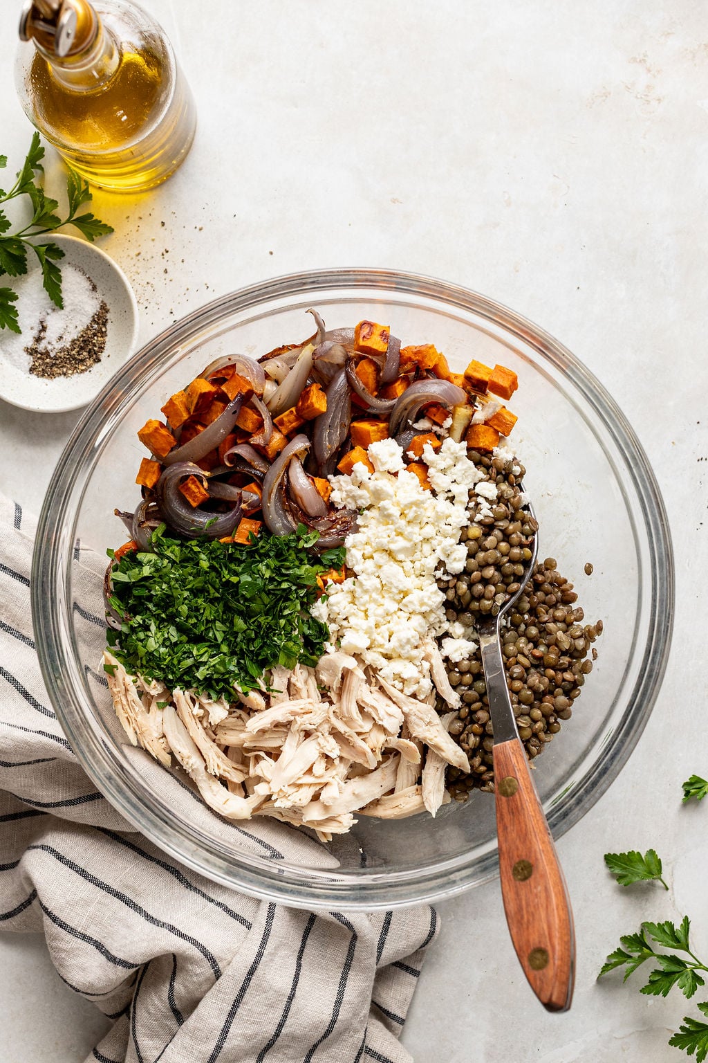 A glass bowl with shredded chicken, cooked lentils, feta cheese, chopped parsley, roasted sweet potato, and roasted red onion, with a fork inside. Olive oil bottle and pepper nearby.