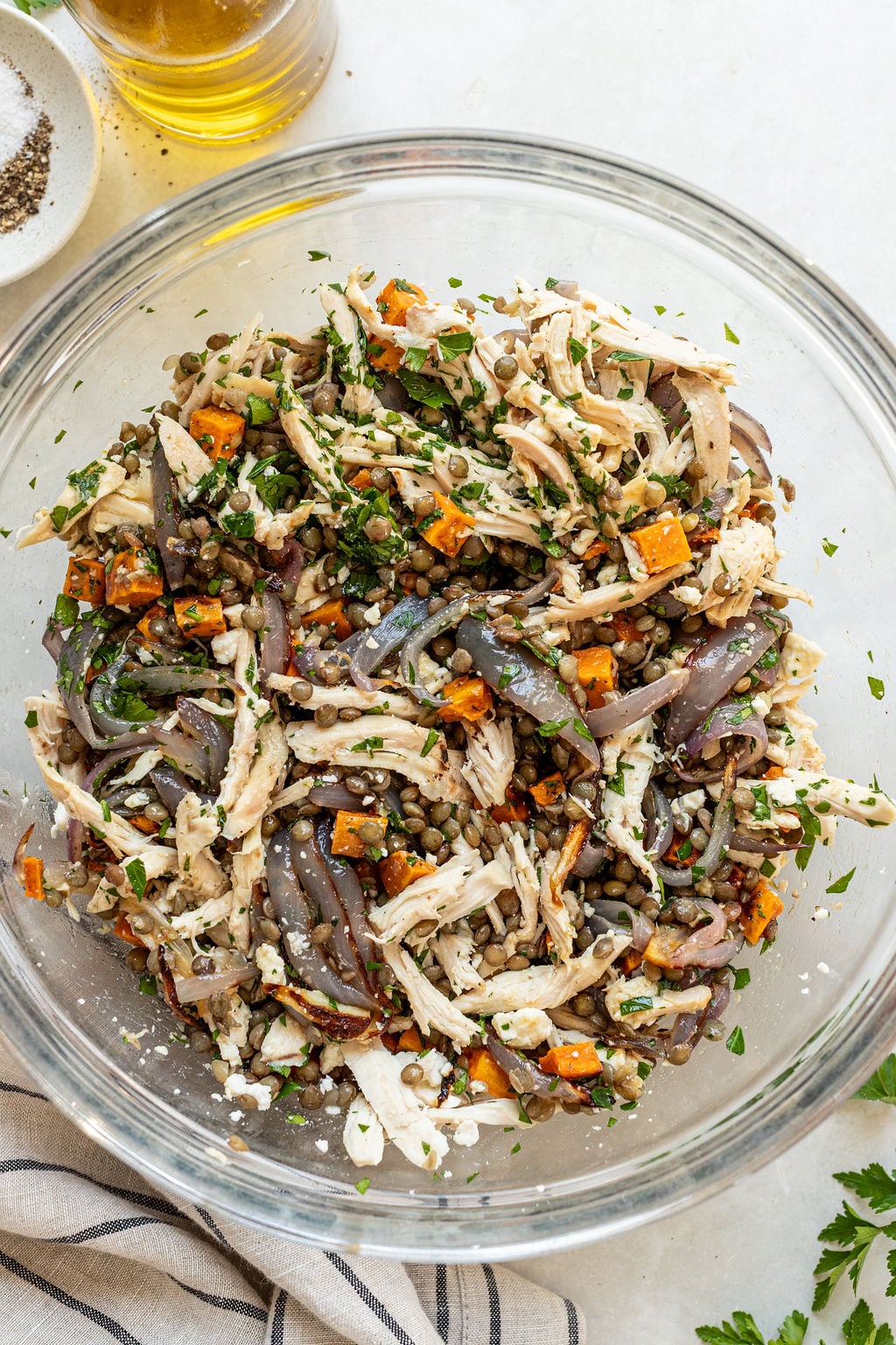 A glass bowl filled with a salad made of shredded chicken, lentils, roasted sweet potatoes, red onions, chopped herbs, and crumbled cheese.