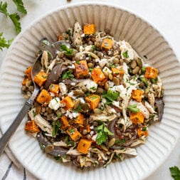 A bowl of salad with roasted sweet potato, shredded chicken, red onions, crumbled cheese, and chopped parsley, with a fork on the side.