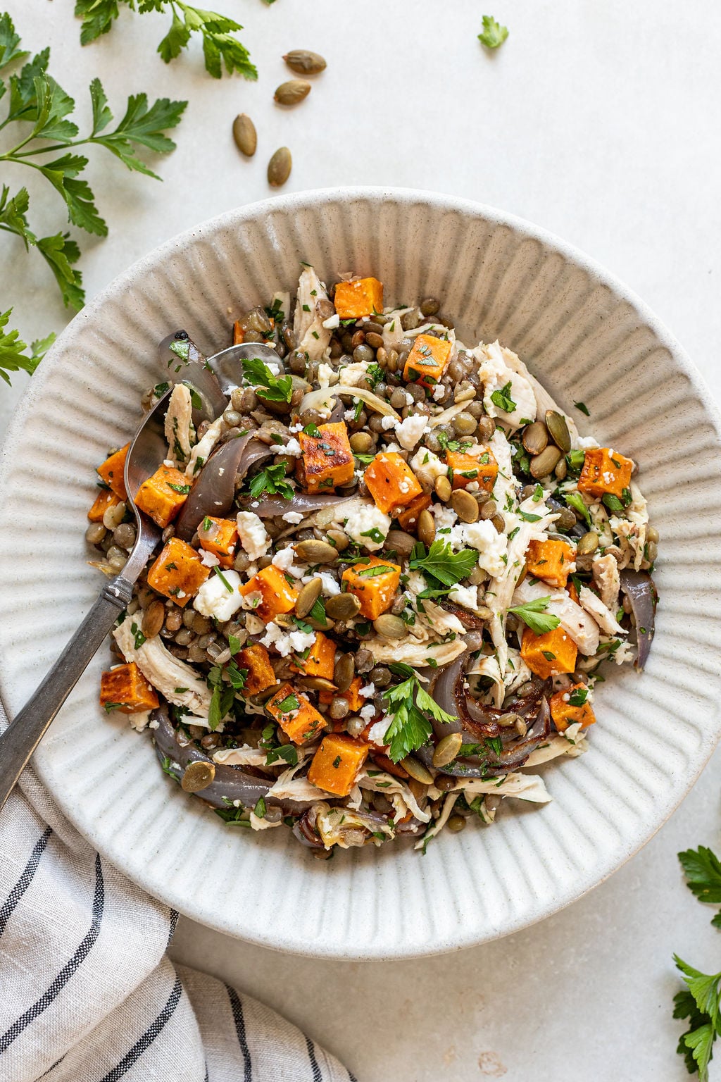 A bowl of salad with roasted sweet potato, shredded chicken, red onions, crumbled cheese, and chopped parsley, with a fork on the side.