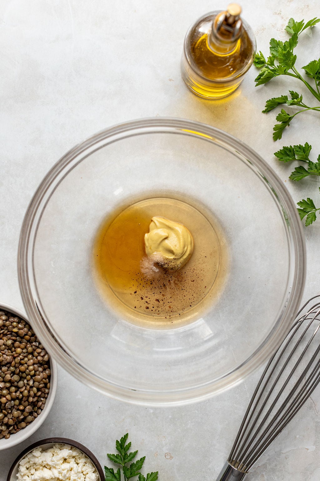 A glass bowl with mustard, vinegar, and pepper sits on a counter, surrounded by olive oil, parsley, cooked lentils, cheese, and a whisk.