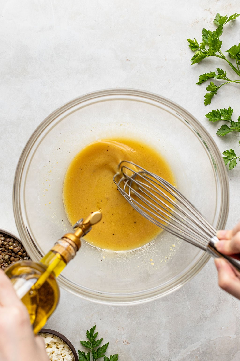 A person whisking vinaigrette in a glass bowl while pouring olive oil in, with fresh herbs and ingredients nearby on a light surface.