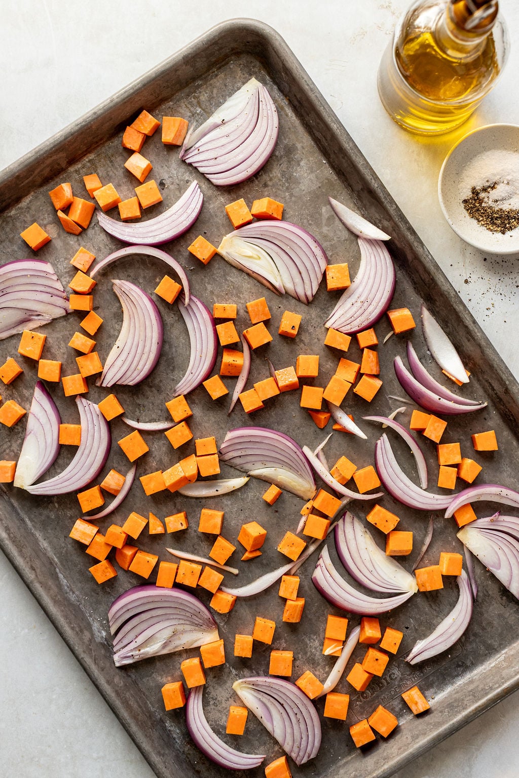 A baking sheet with sliced red onions and diced sweet potatoes arranged in a single layer, next to a bowl of seasoning and a bottle of olive oil.