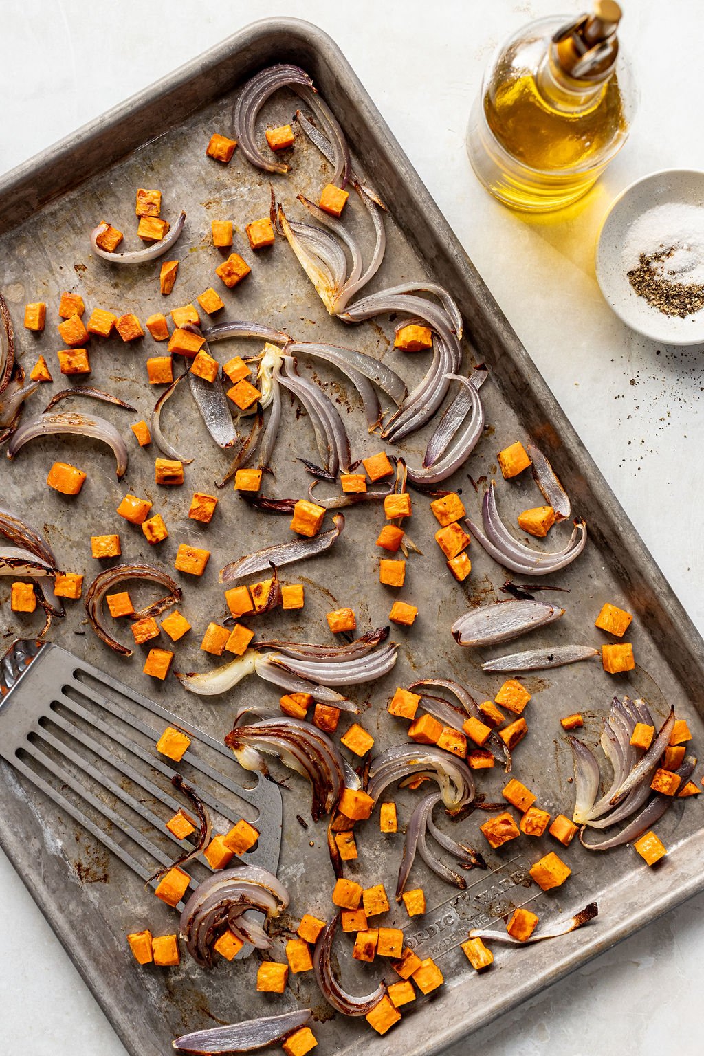 Baking sheet with roasted sweet potato cubes and red onion slices, a metal spatula, a bottle of olive oil, and a small bowl of salt and pepper on the side.