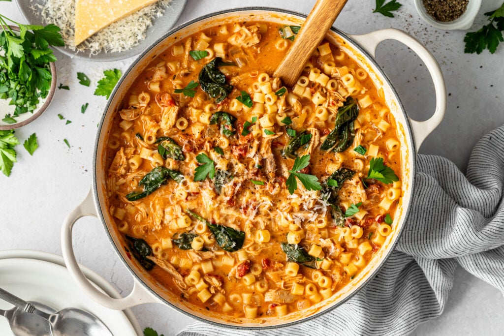 A pot of creamy tomato pasta soup with ditalini, spinach, shredded chicken, and parsley garnish, with a wooden spoon for serving.