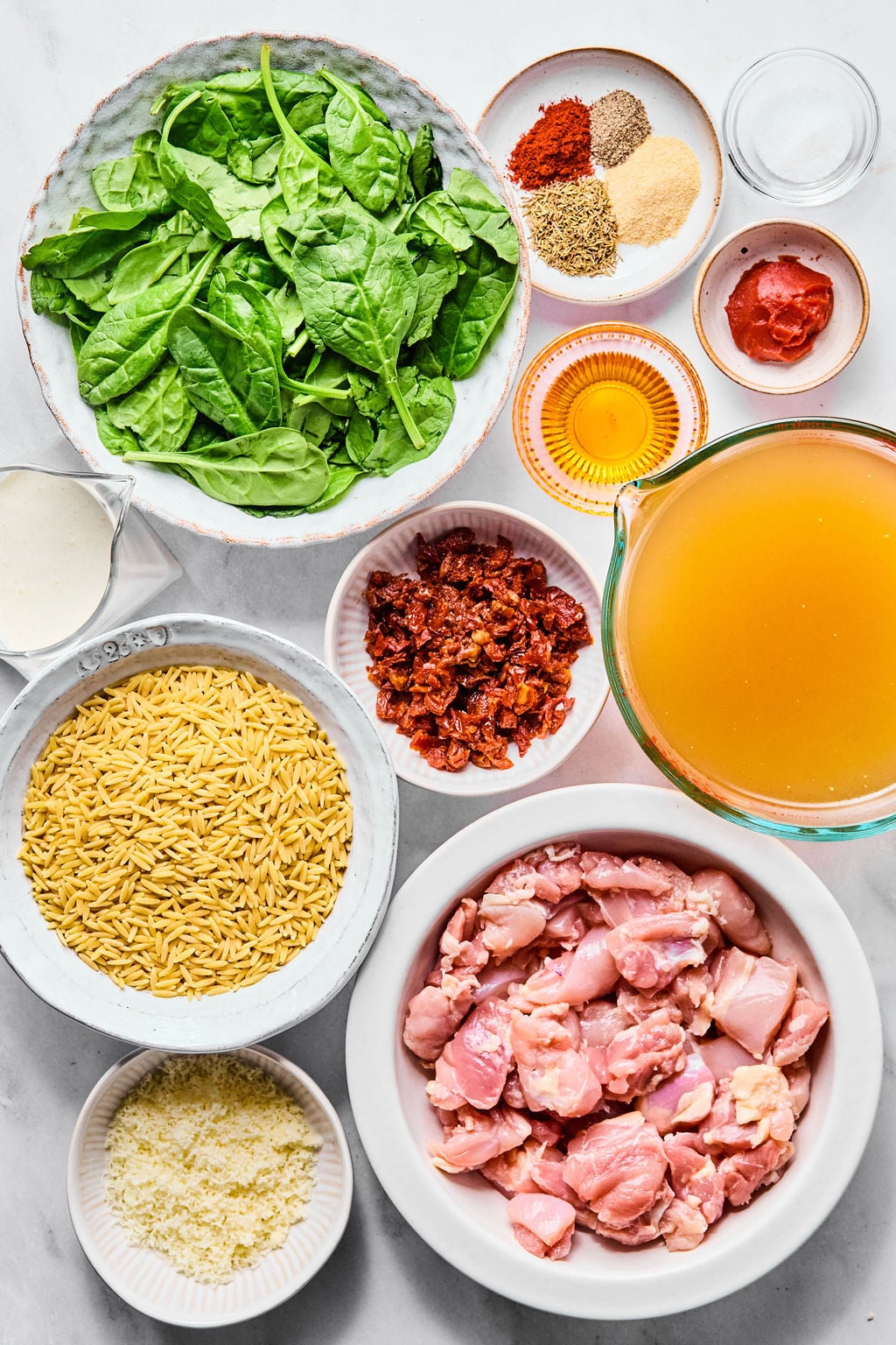 A flat lay of ingredients including spinach, seasonings, tomato paste, cream, sun-dried tomatoes, chicken broth, orzo pasta, raw chicken pieces, and grated cheese in separate bowls.