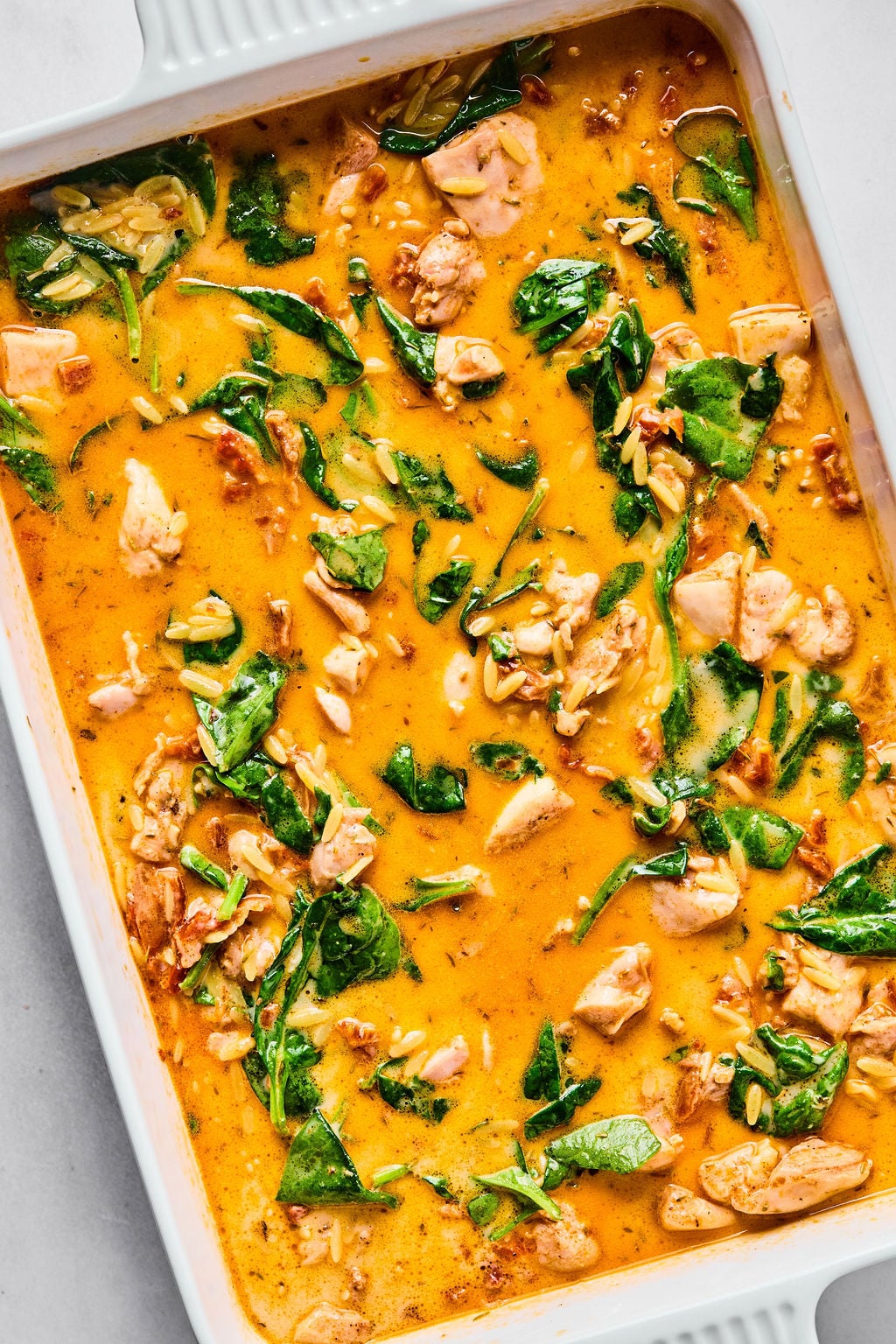 Baked chicken in a creamy orange sauce with spinach and sun-dried tomatoes in a white rectangular baking dish.