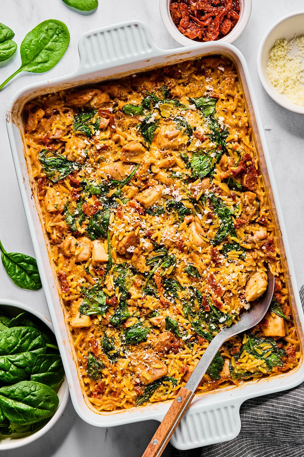 A baked casserole dish filled with a creamy mixture of chicken, spinach, sundried tomatoes, and rice, with grated cheese sprinkled on top. A serving spoon rests inside the dish.