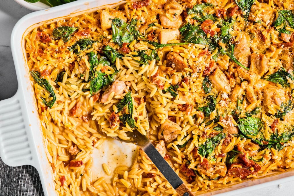 A baked orzo casserole with spinach, sun-dried tomatoes, and chunks of chicken, partially served in a white baking dish with a serving spoon.