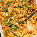 Baked orzo casserole with visible pieces of chicken, spinach, and sun-dried tomatoes in a creamy sauce, partially served with a metal spoon in a white baking dish.
