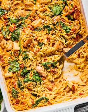 Baked orzo casserole with visible pieces of chicken, spinach, and sun-dried tomatoes in a creamy sauce, partially served with a metal spoon in a white baking dish.