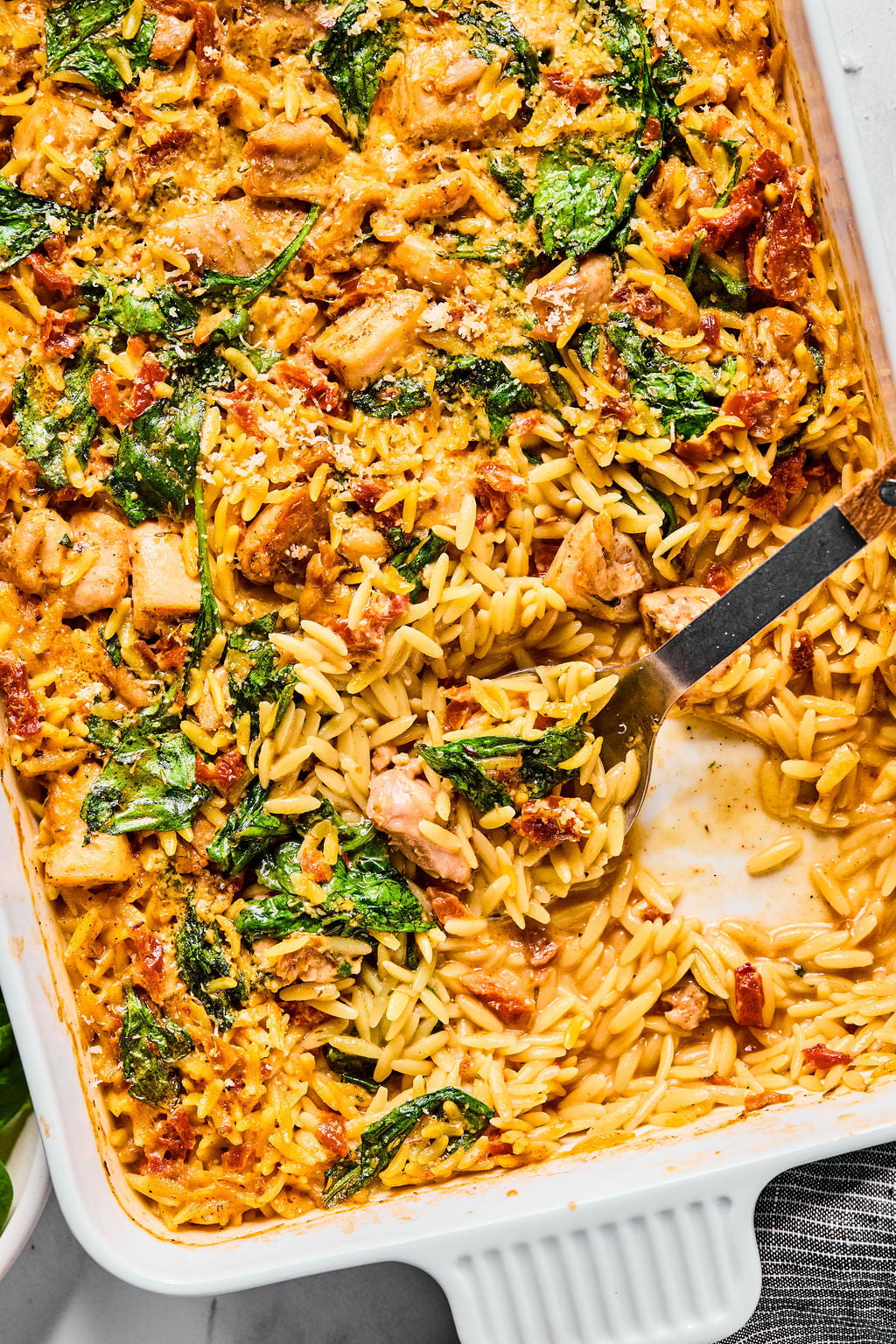 Baked orzo casserole with visible pieces of chicken, spinach, and sun-dried tomatoes in a creamy sauce, partially served with a metal spoon in a white baking dish.
