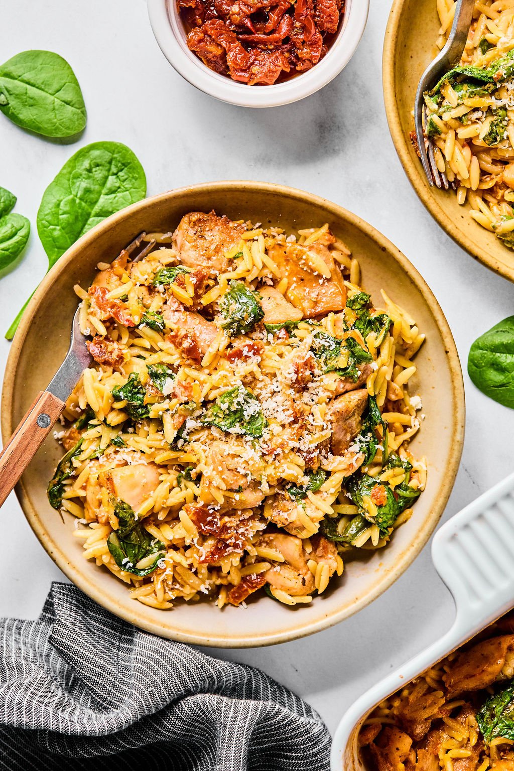 A bowl of orzo pasta with pieces of chicken, spinach, sun-dried tomatoes, and grated cheese, served with a fork on the side.