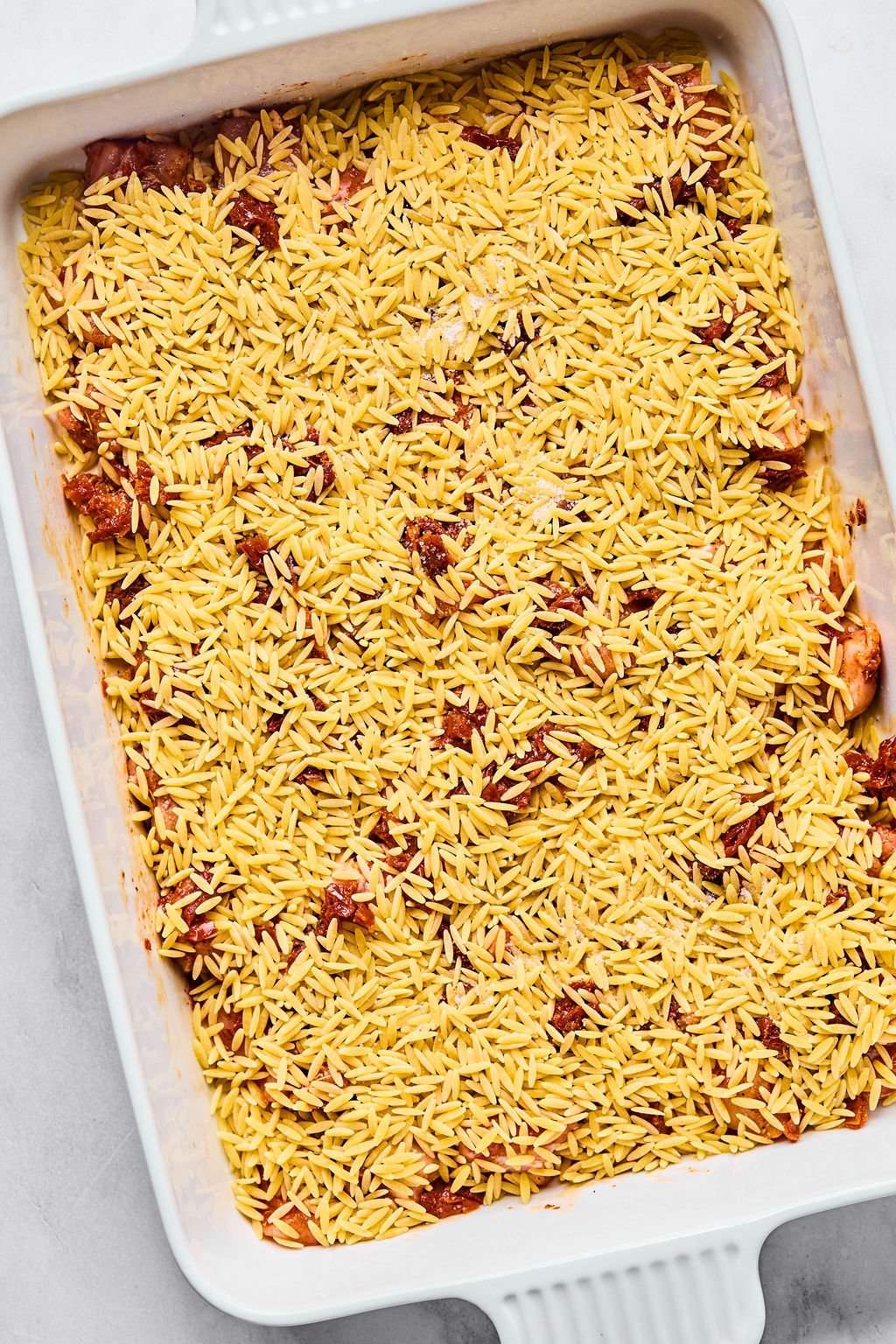 Uncooked orzo pasta mixed with pieces of sun-dried tomatoes spread evenly in a white rectangular baking dish.