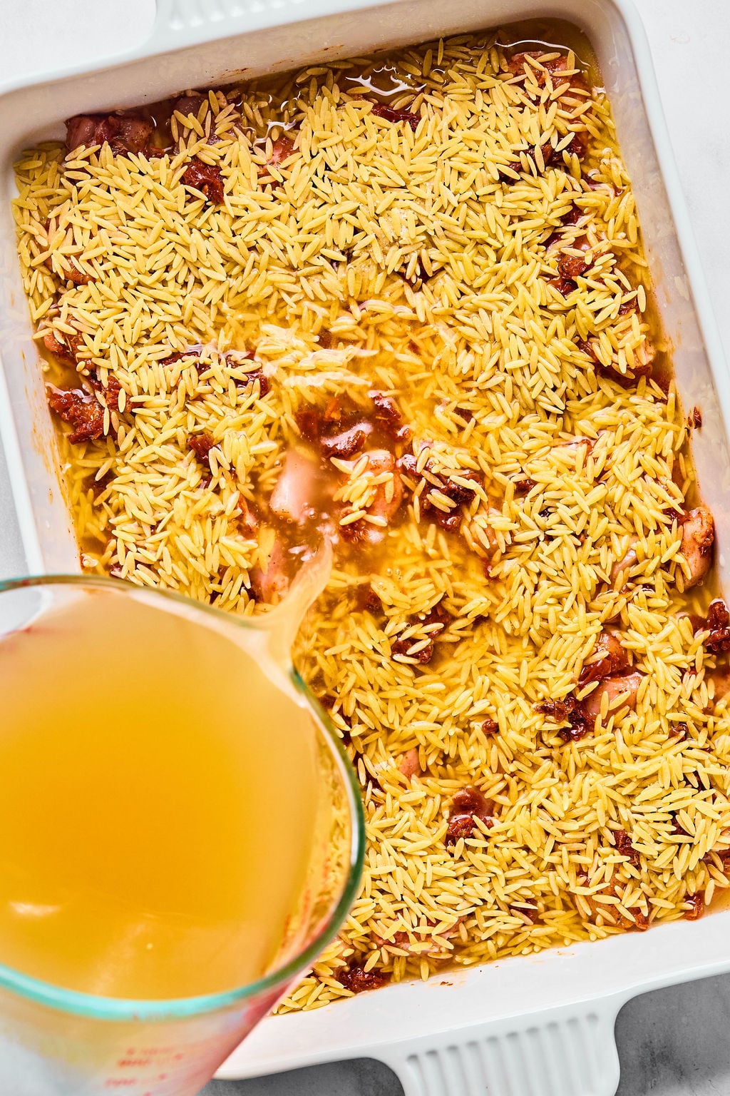 A measuring cup pours broth over uncooked rice and pieces of chicken in a white baking dish.
