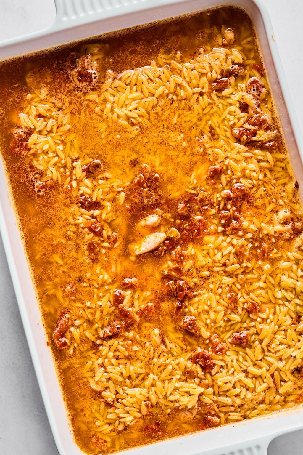 A white baking dish filled with cooked orzo pasta in a tomato-based sauce, with visible pieces of tomato and broth.