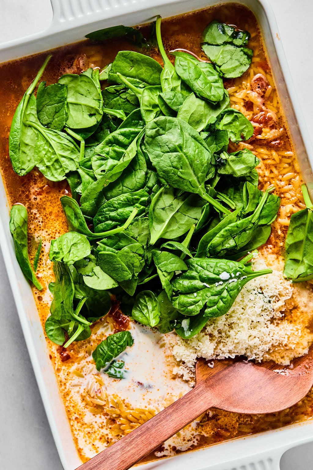 A baking dish filled with uncooked pasta, sauce, fresh spinach leaves, grated cheese, and a wooden spoon.