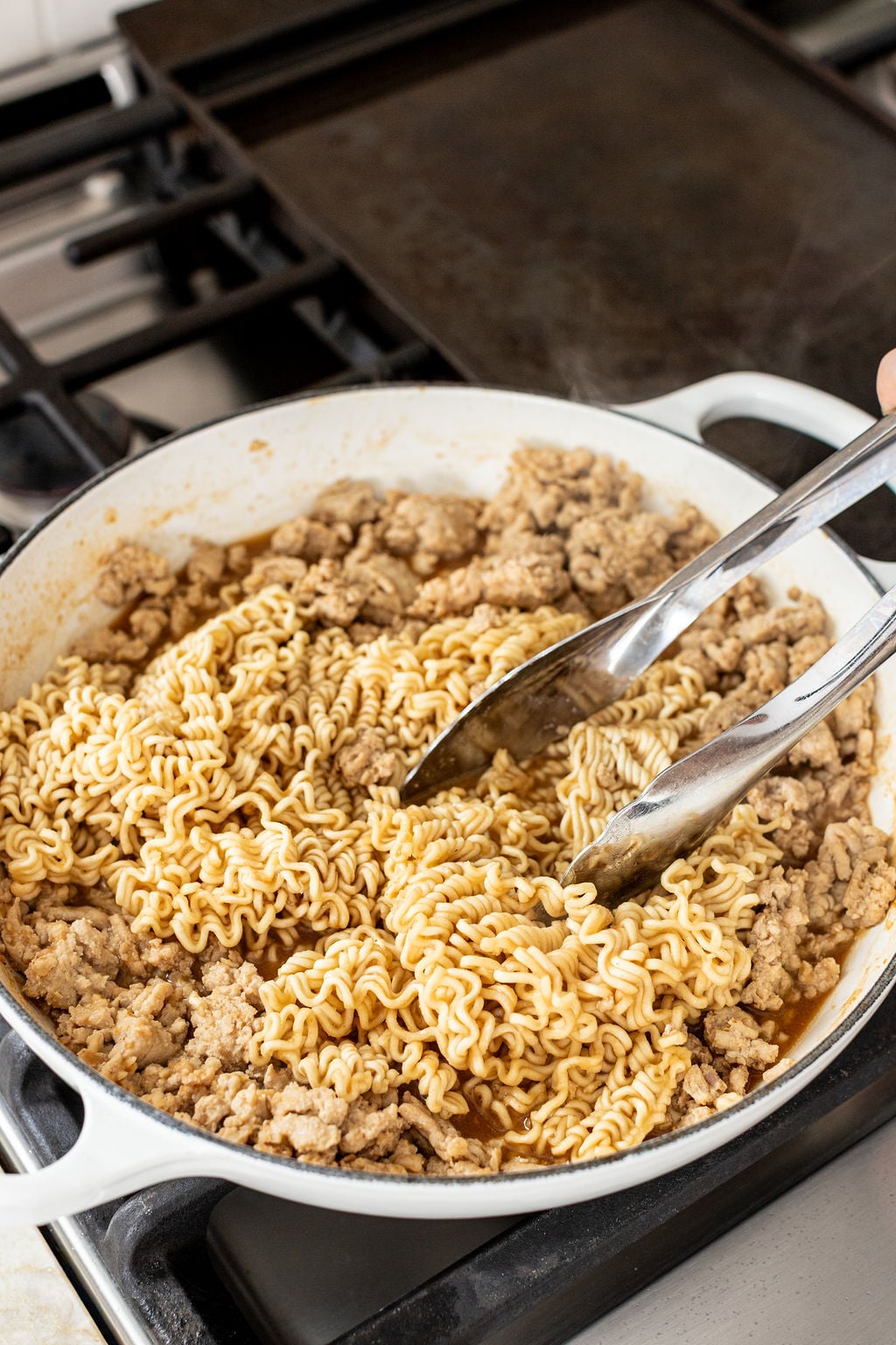 A pan on a stovetop contains uncooked instant ramen noodles and browned ground meat being mixed with metal tongs.