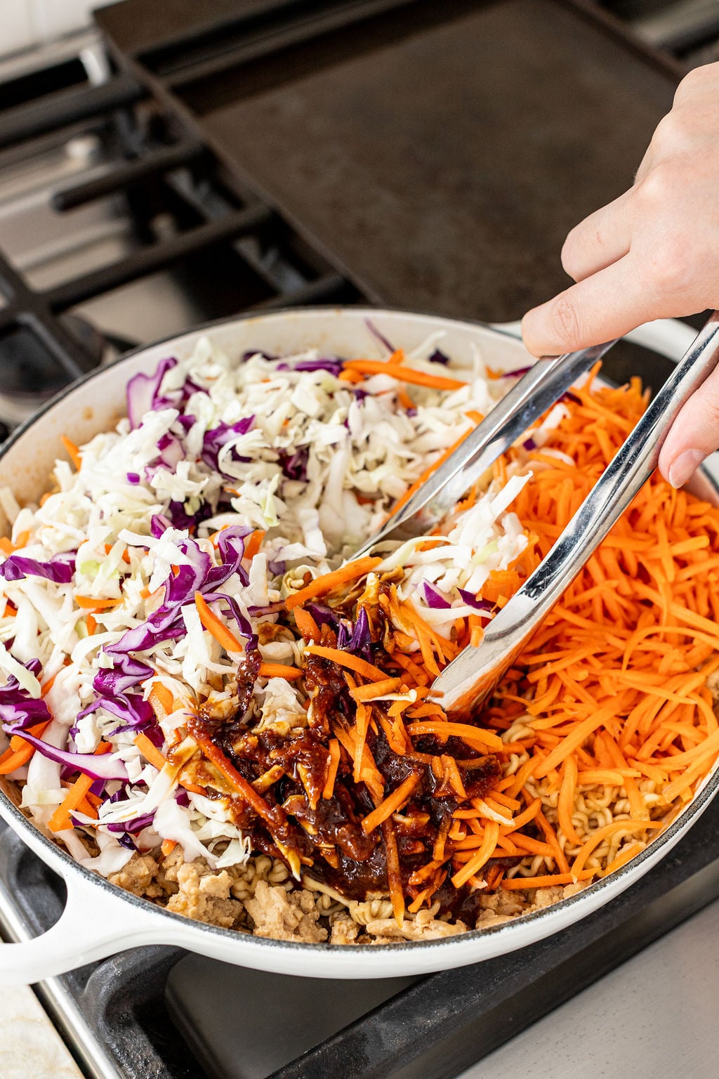 A person uses tongs to mix shredded cabbage, carrots, ground meat, and sauce in a white skillet on a stovetop.