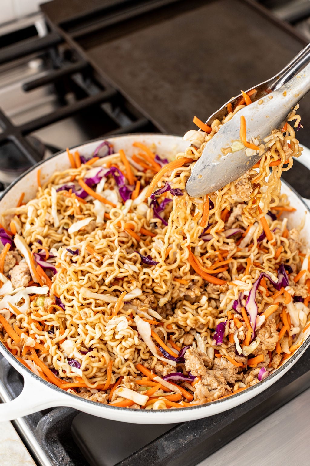 A skillet on a stove containing cooked ramen noodles, ground meat, shredded carrots, and cabbage being stirred with metal tongs.