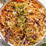 A large pan filled with cooked ramen noodles, shredded carrots, cabbage, green onions, and chopped peanuts, with metal tongs resting on the side.