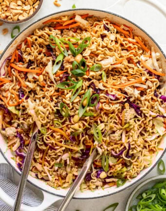A large pan filled with cooked ramen noodles, shredded carrots, cabbage, green onions, and chopped peanuts, with metal tongs resting on the side.