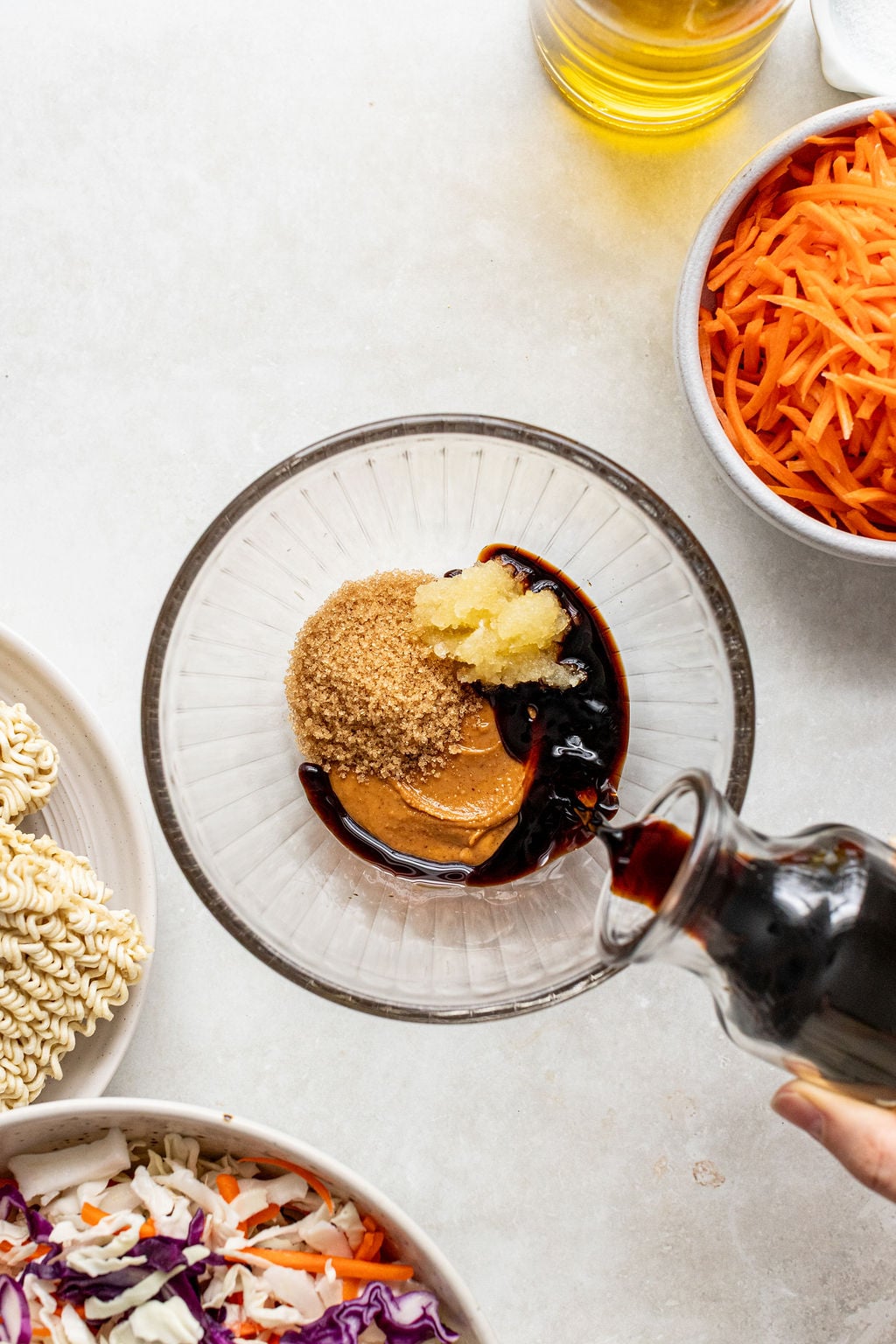 A glass bowl with brown sugar, peanut butter, grated garlic, and dark sauce being poured in, surrounded by shredded carrots, dry ramen noodles, and chopped cabbage.