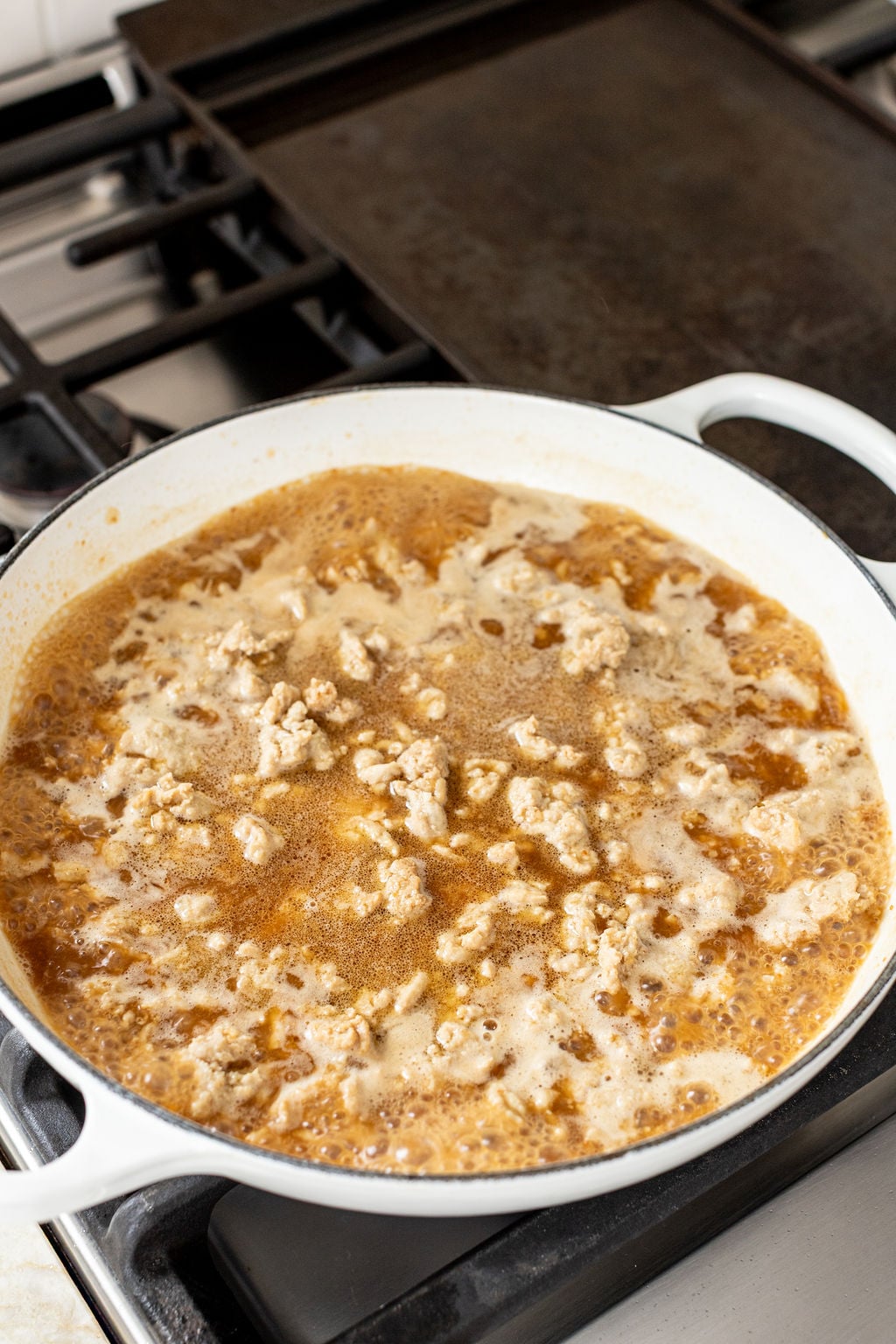 Ground meat simmering in a broth inside a white pot on a stovetop, with steam and bubbles visible on the surface.
