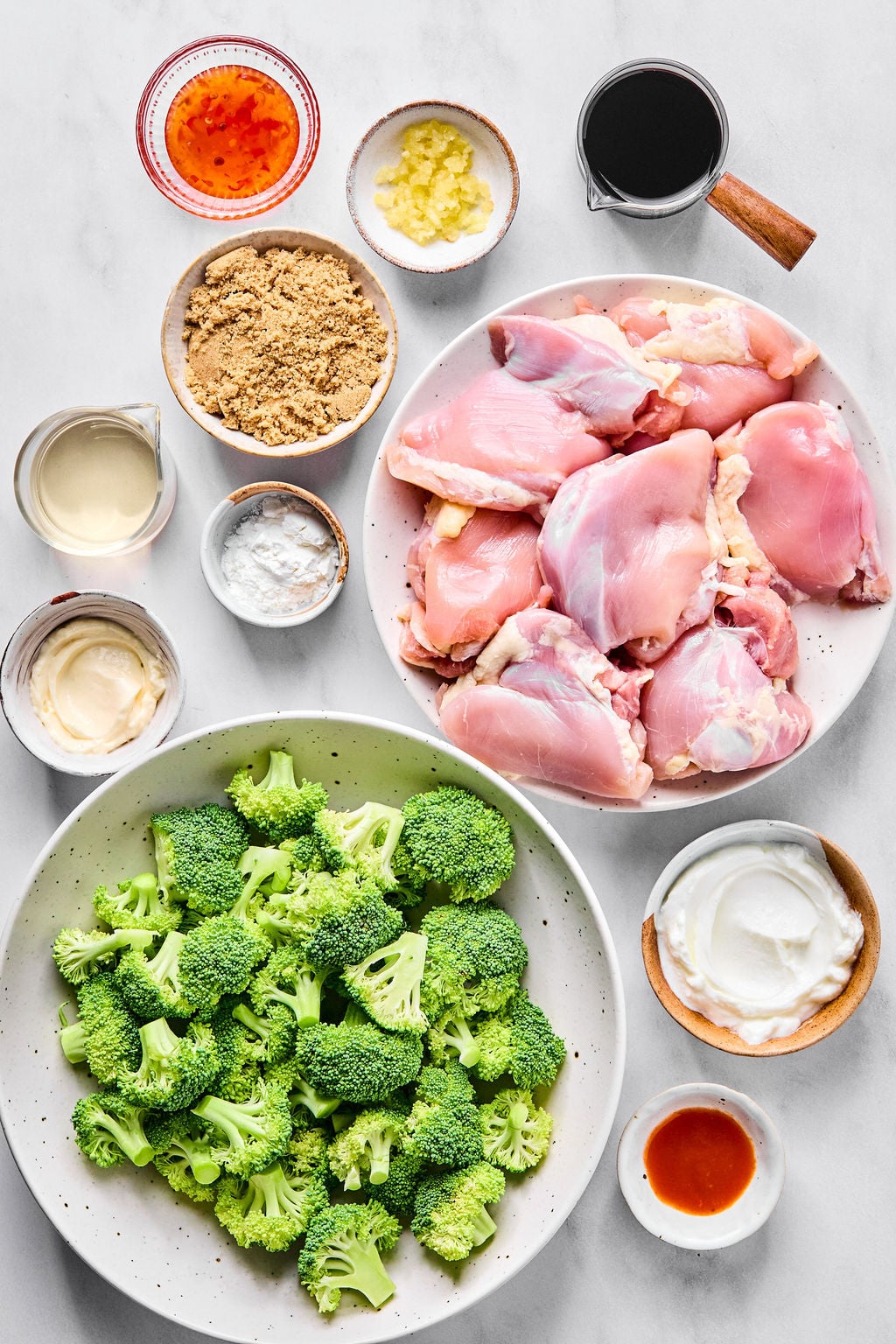 Bowls containing raw chicken thighs, chopped broccoli, brown sugar, garlic, sauces, and seasonings are arranged on a white surface, ready for meal preparation.