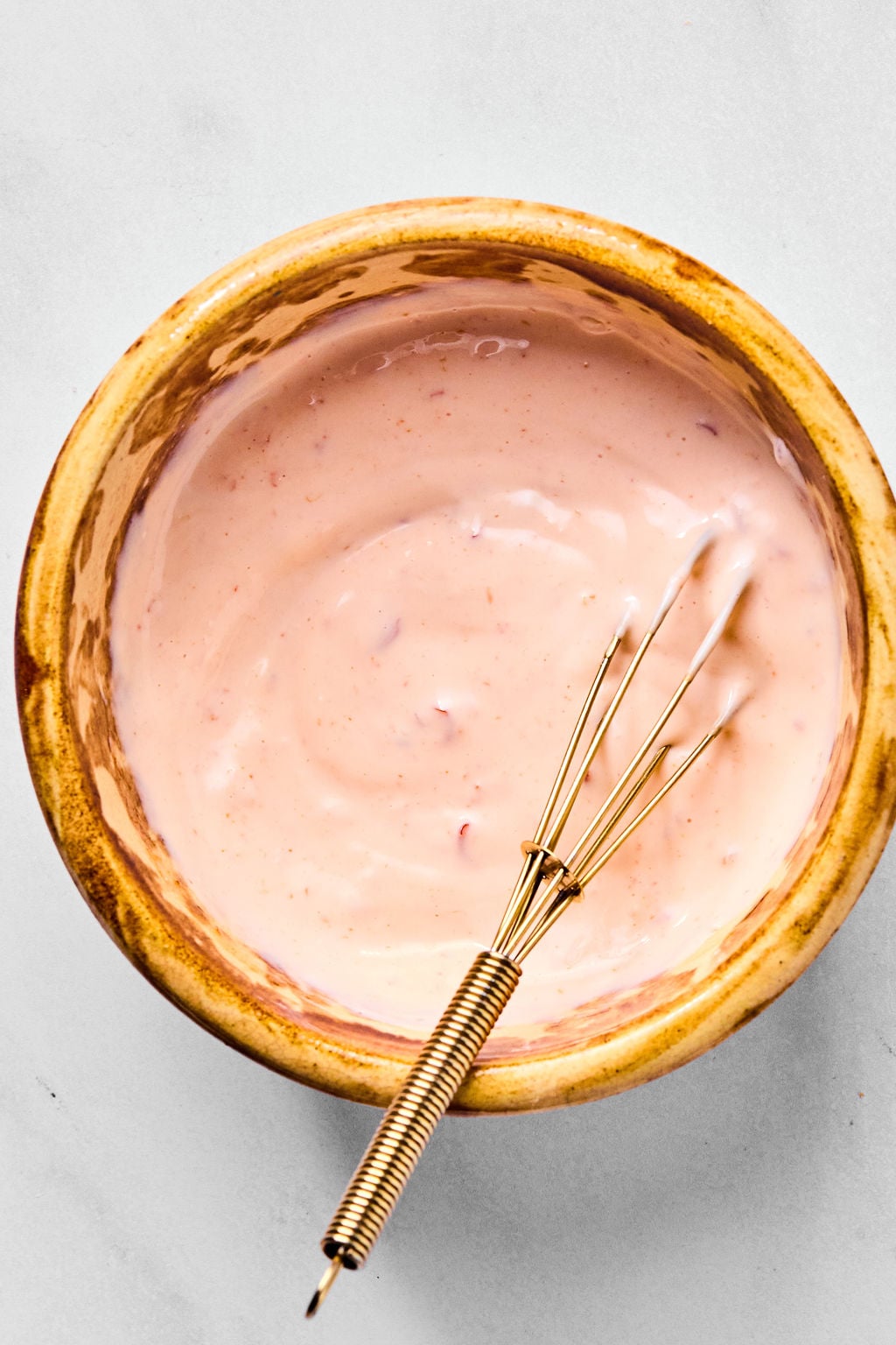 A small bowl of pink sauce with a metal whisk resting inside, placed on a light-colored surface.