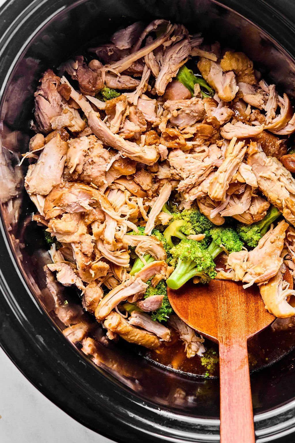 Shredded cooked chicken and broccoli in a slow cooker with a wooden spoon.