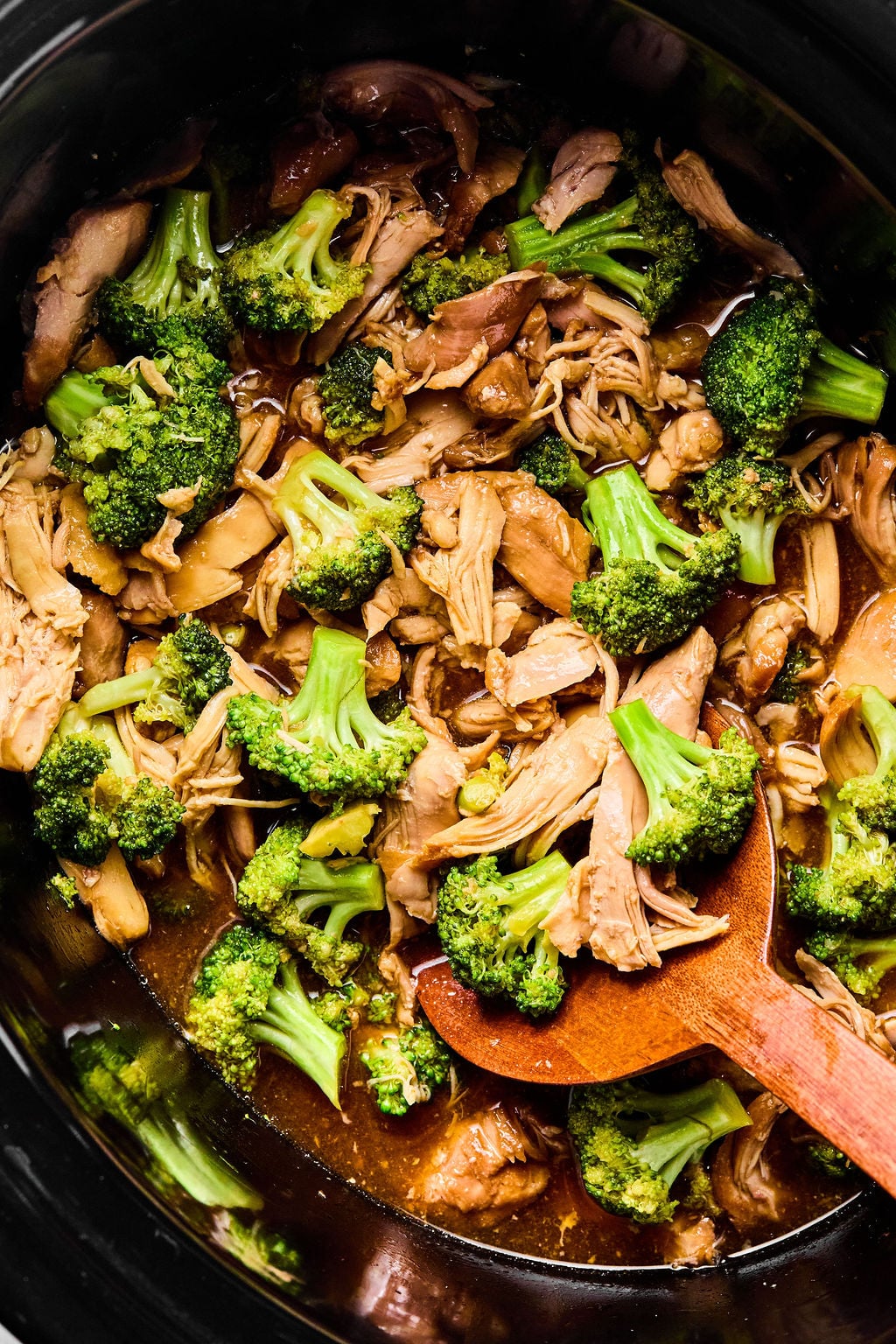 Chicken and broccoli cooked in a savory sauce inside a slow cooker, with a wooden spoon stirring the mixture.
