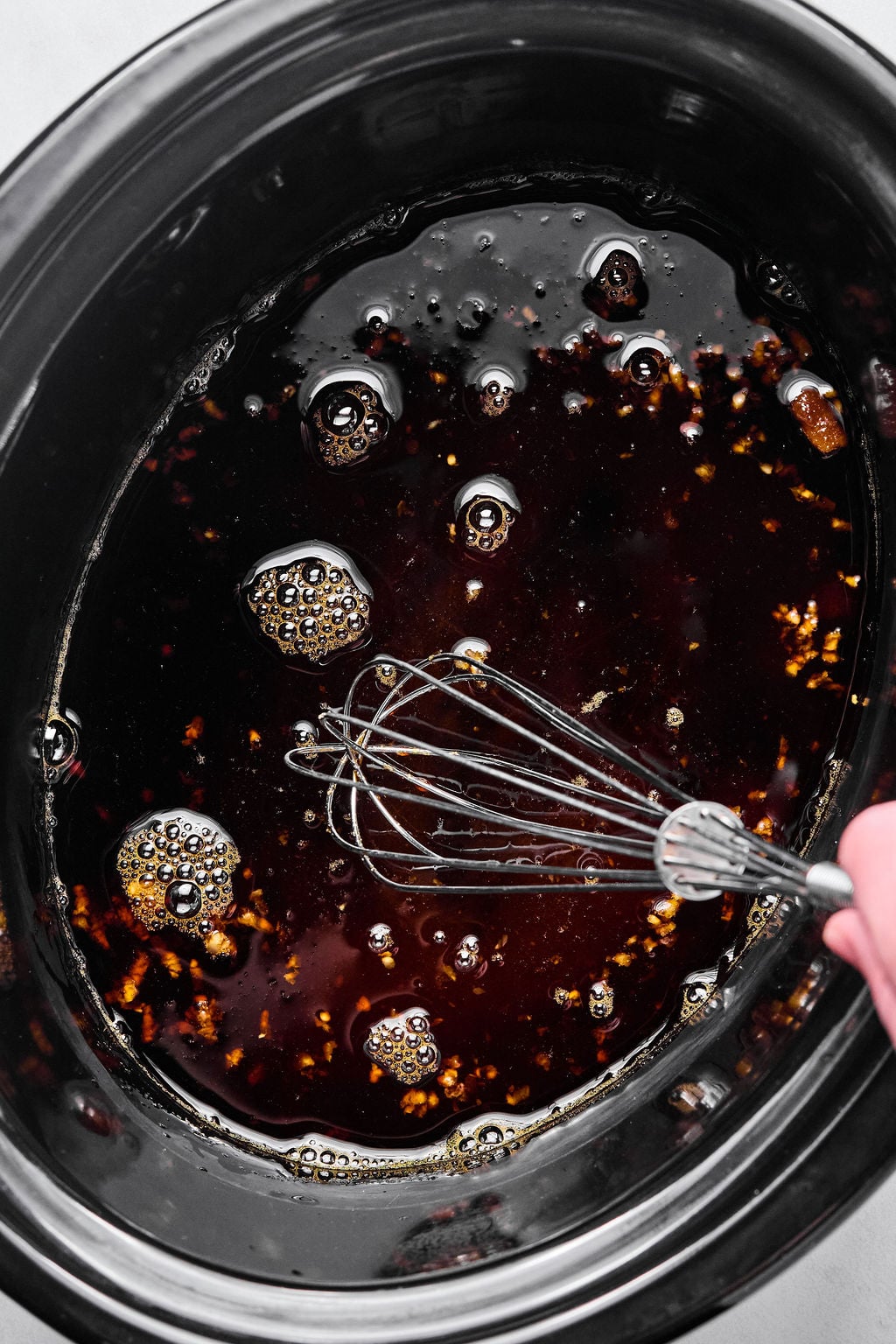 A hand holding a whisk stirs a dark liquid with bubbles and small solid pieces in a black slow cooker.