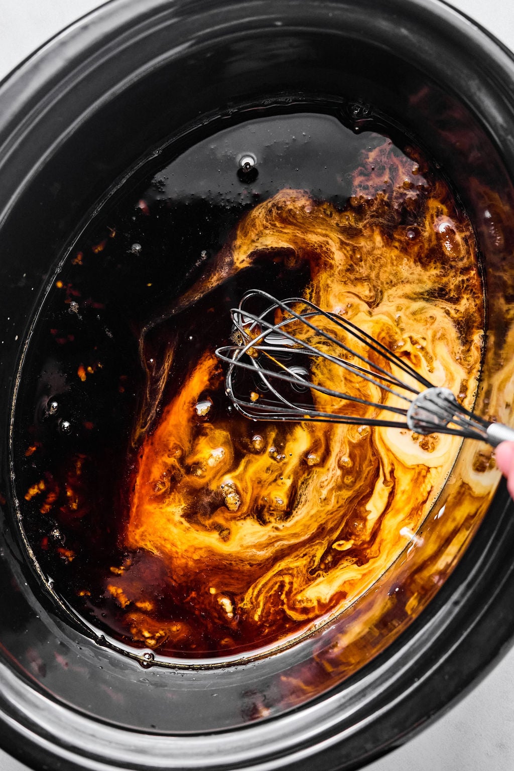 A hand uses a metal whisk to mix dark and light liquids in a black slow cooker.