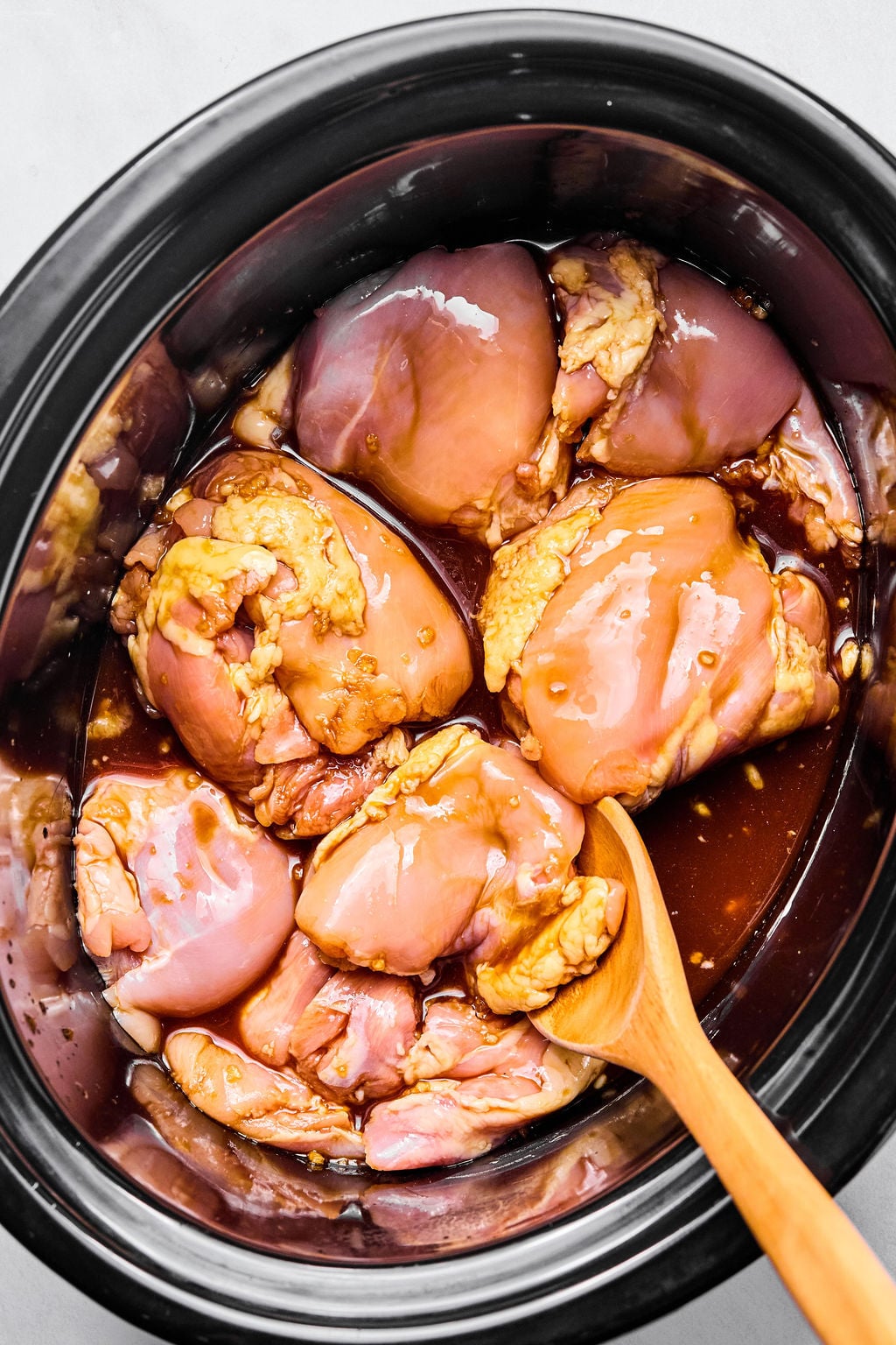 Raw chicken thighs marinating in a brown sauce inside a slow cooker, with a wooden spoon resting on the side.