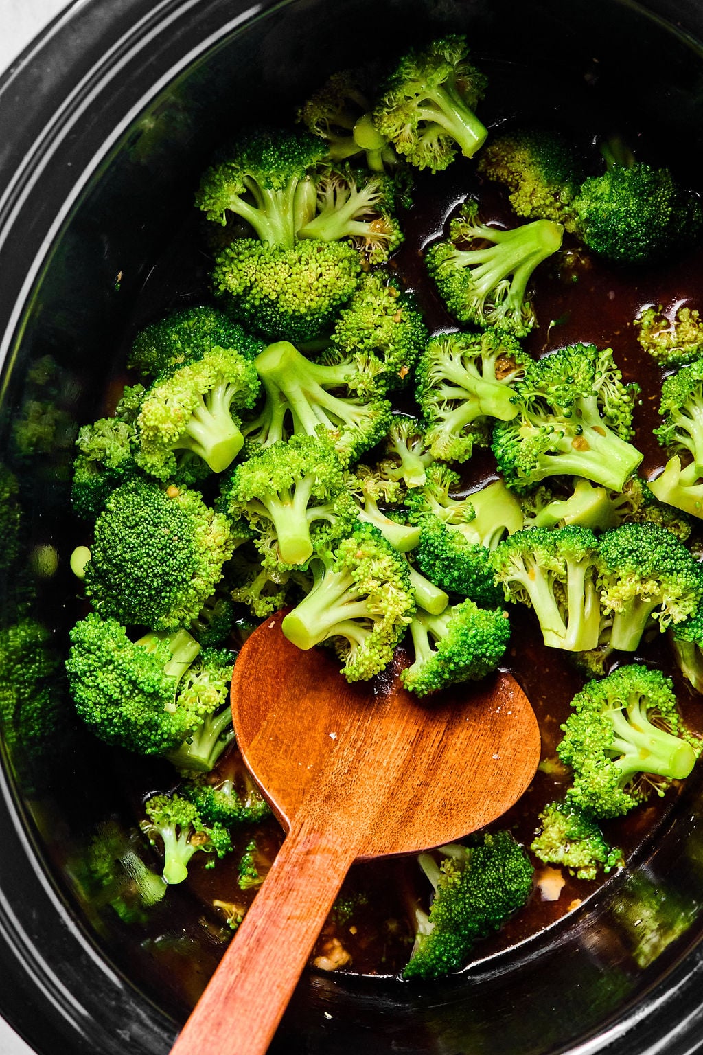 Broccoli florets in a black slow cooker with a wooden spoon, sitting on top of a dark sauce.