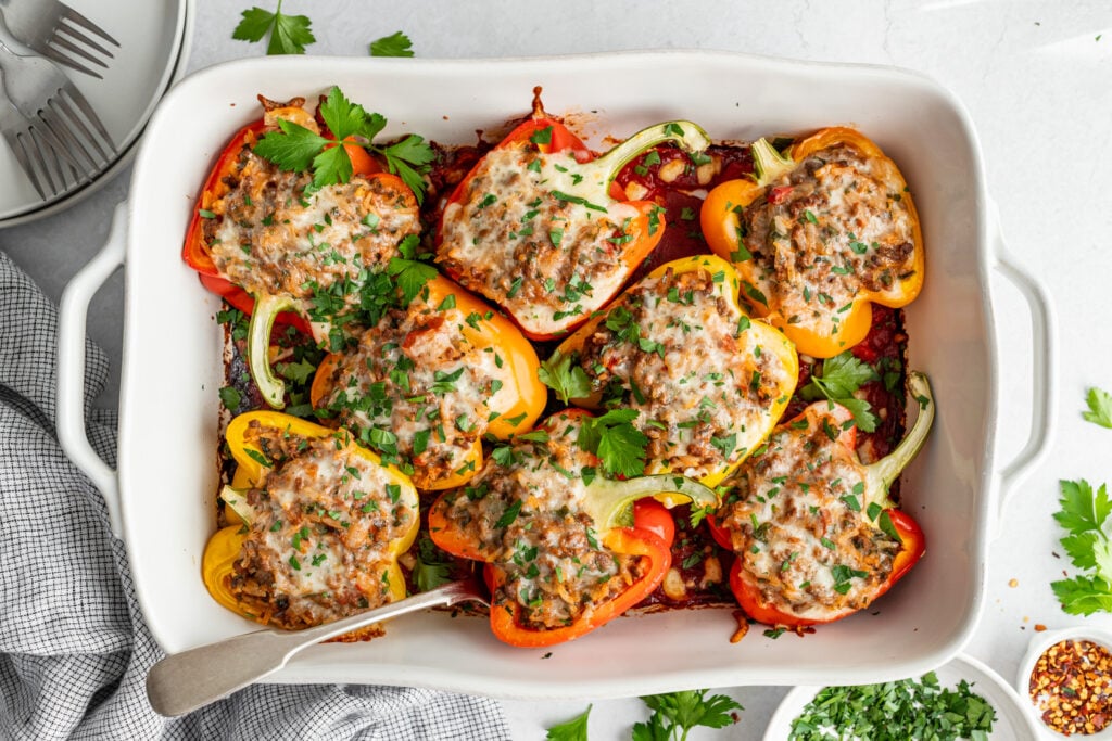 A white baking dish filled with halved bell peppers stuffed with ground meat and topped with melted cheese and chopped parsley.